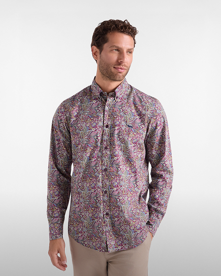 Un uomo con capelli ricci castani e barba corta indossa una camicia a maniche lunghe con fantasia paisley multicolore nei toni del viola, rosa e verde, abbinata a pantaloni beige.