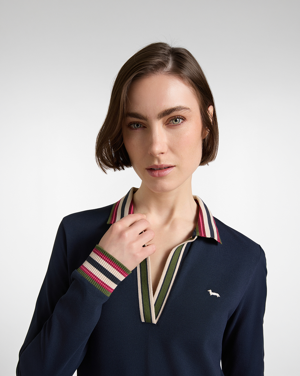 Immagine di campagna per la collezione FW26 Harmont & Blaine, focalizzata sulla polo per lei. Una modella con taglio bob castano chiaro &egrave; ritratta a mezzo busto su sfondo neutro mentre indossa una maglia stile polo a maniche lunghe blu scuro. Il capo presenta uno scollo a V profondo, colletto e polsini a costine con righe multicolor (rosa, verde, crema e nero) e l'iconico logo del bassotto ricamato in bianco sul petto.