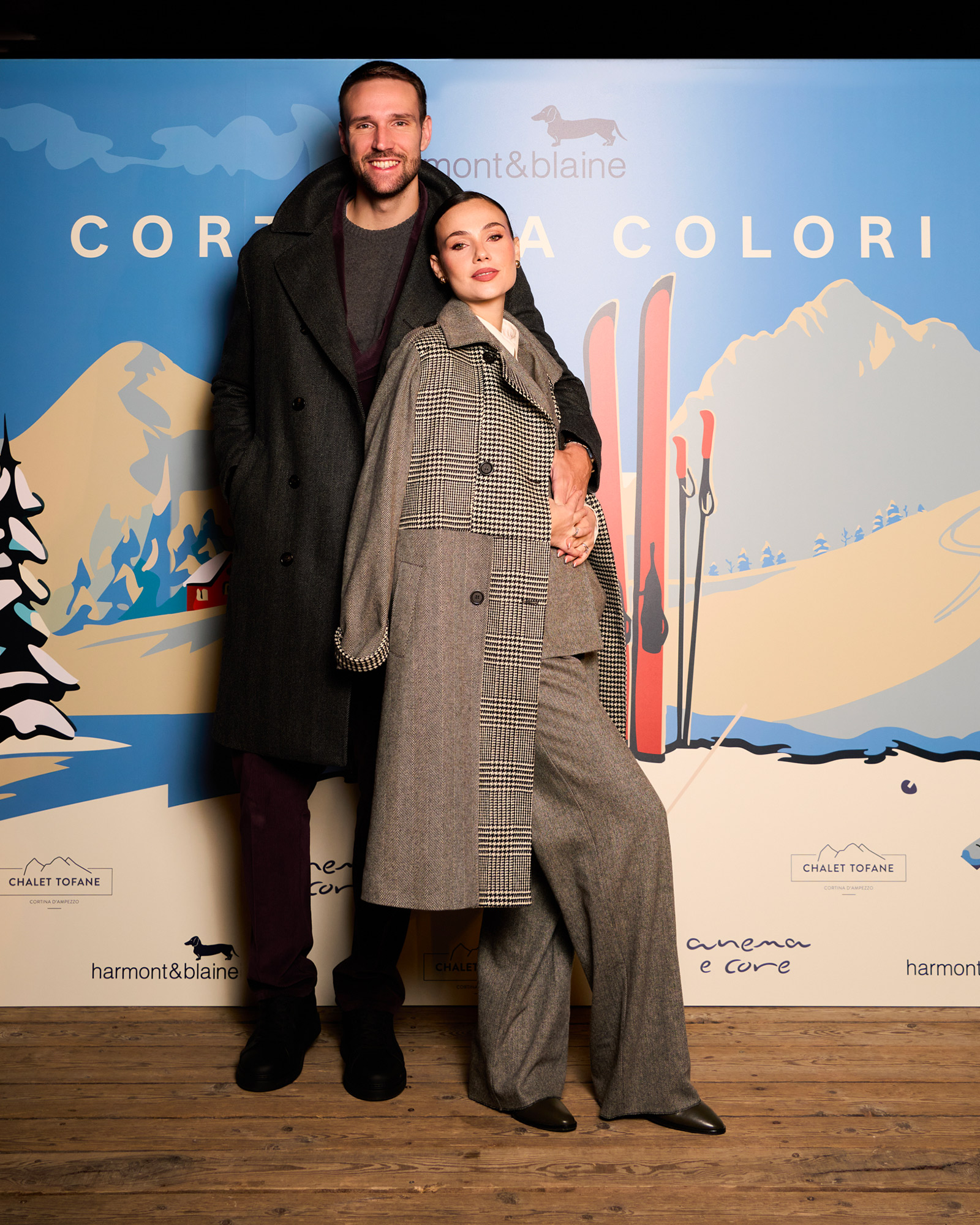 Woman in a grey checked coat and tailored suit with a man in a dark coat in front of the Cortina a Colori backdrop.