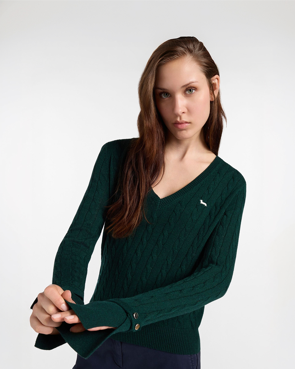 Immagine di campagna per la collezione FW26 Harmont & Blaine, focalizzata sulla maglieria donna. Una modella con lunghi capelli castani &egrave; ritratta a mezzo busto su sfondo neutro mentre indossa un maglione con scollo a V in maglia a trecce color verde bosco. Sul petto &egrave; ricamato il logo iconico del bassotto in grigio, mentre le mani in primo piano evidenziano i polsini rovesciati rifiniti con due bottoni decorativi dorati.