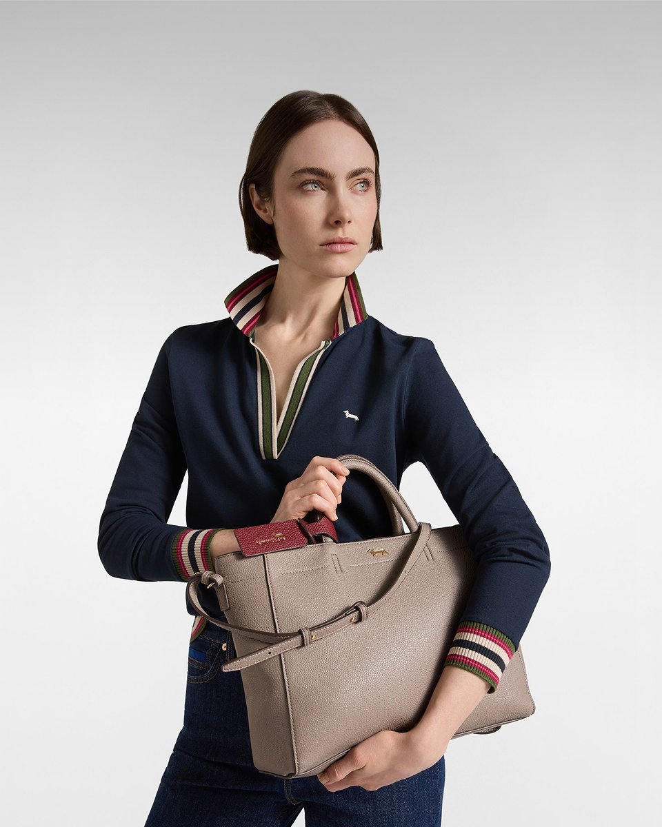 Una donna con capelli corti castani indossa una polo a maniche lunghe blu scuro con scollo a V e dettagli a righe e tiene in braccio una borsa tote in pelle martellata grigio tortora con manici rigidi e tracolla.
