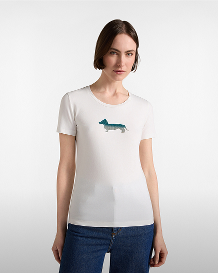 Una donna con capelli corti castani indossa una t-shirt bianca a maniche corte con un bassotto sfumato di azzurro e verde stampato al centro del petto, abbinata a jeans blu scuro.