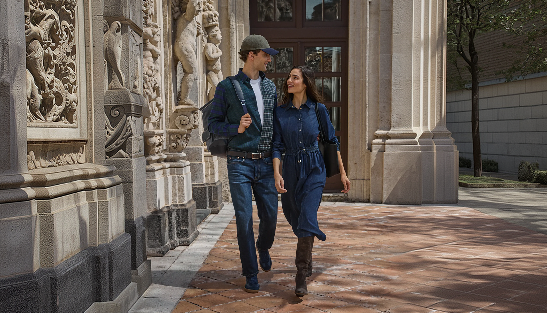 Immagine promozionale per la collezione Autunno/Inverno 2026 raffigurante un uomo e una donna che camminano sorridenti affiancati lungo un porticato storico in pietra; l'uomo indossa un cardigan a quadri verde e blu scuro su t-shirt bianca, jeans e cappellino con visiera verde oliva, mentre la donna indossa uno chemisier lungo in denim blu scuro cinturato in vita, stivali alti in camoscio marrone e porta una borsa a spalla nera.