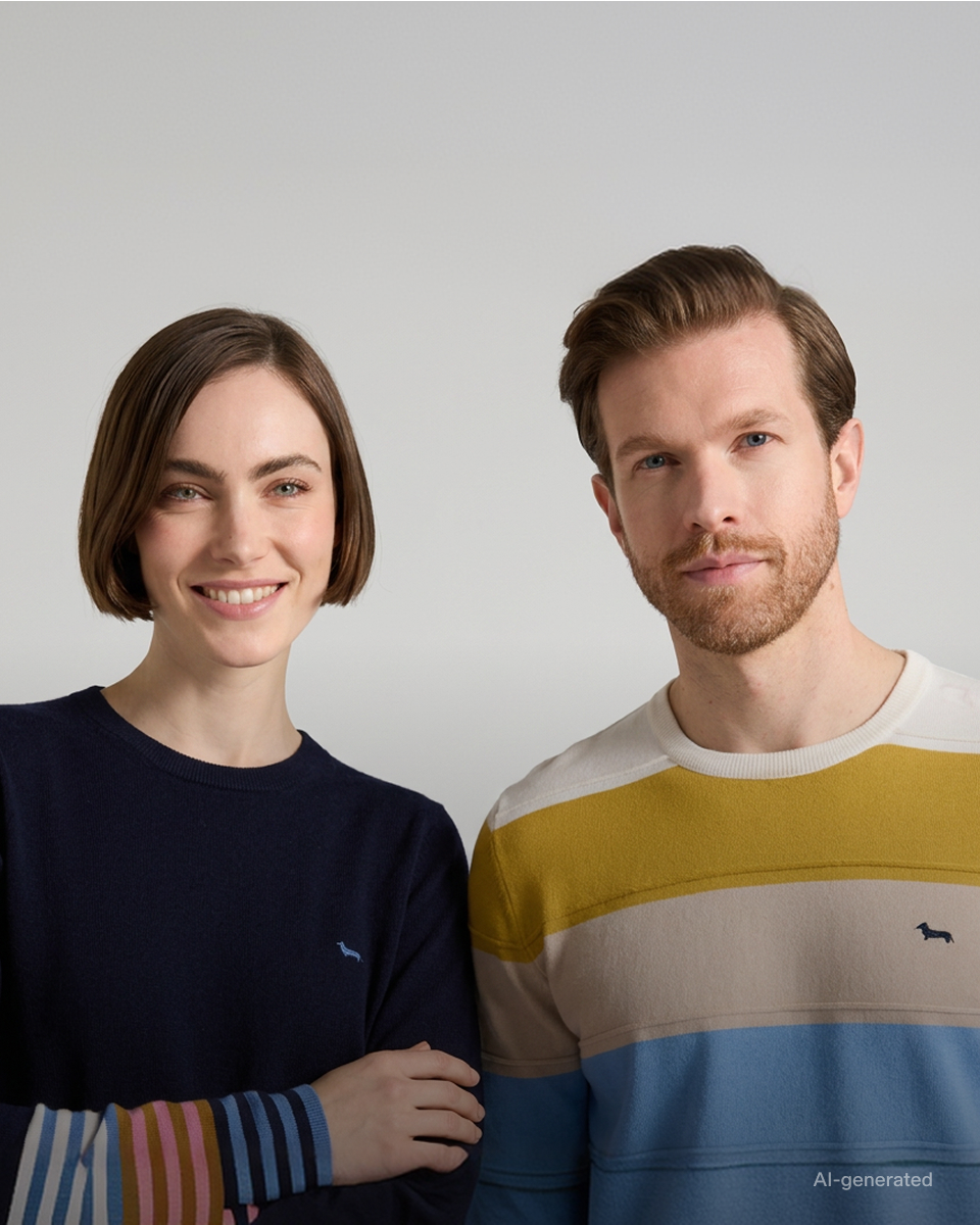 Immagine di campagna generata con l'intelligenza artificiale per la collezione FW26, a tema "Righe per Lei e per Lui". La foto ritrae a mezzo busto una donna e un uomo affiancati su uno sfondo grigio neutro. La donna a sinistra, con un taglio corto castano e sorridente, indossa una maglia girocollo blu scuro caratterizzata da polsini a righe multicolore nei toni del rosa, azzurro e ocra, con un piccolo logo ricamato sul petto. L'uomo a destra, con barba e capelli castani, sfoggia un maglione girocollo a ampie righe orizzontali ocra, panna e azzurro, con il logo del bassotto ricamato al centro del torace.