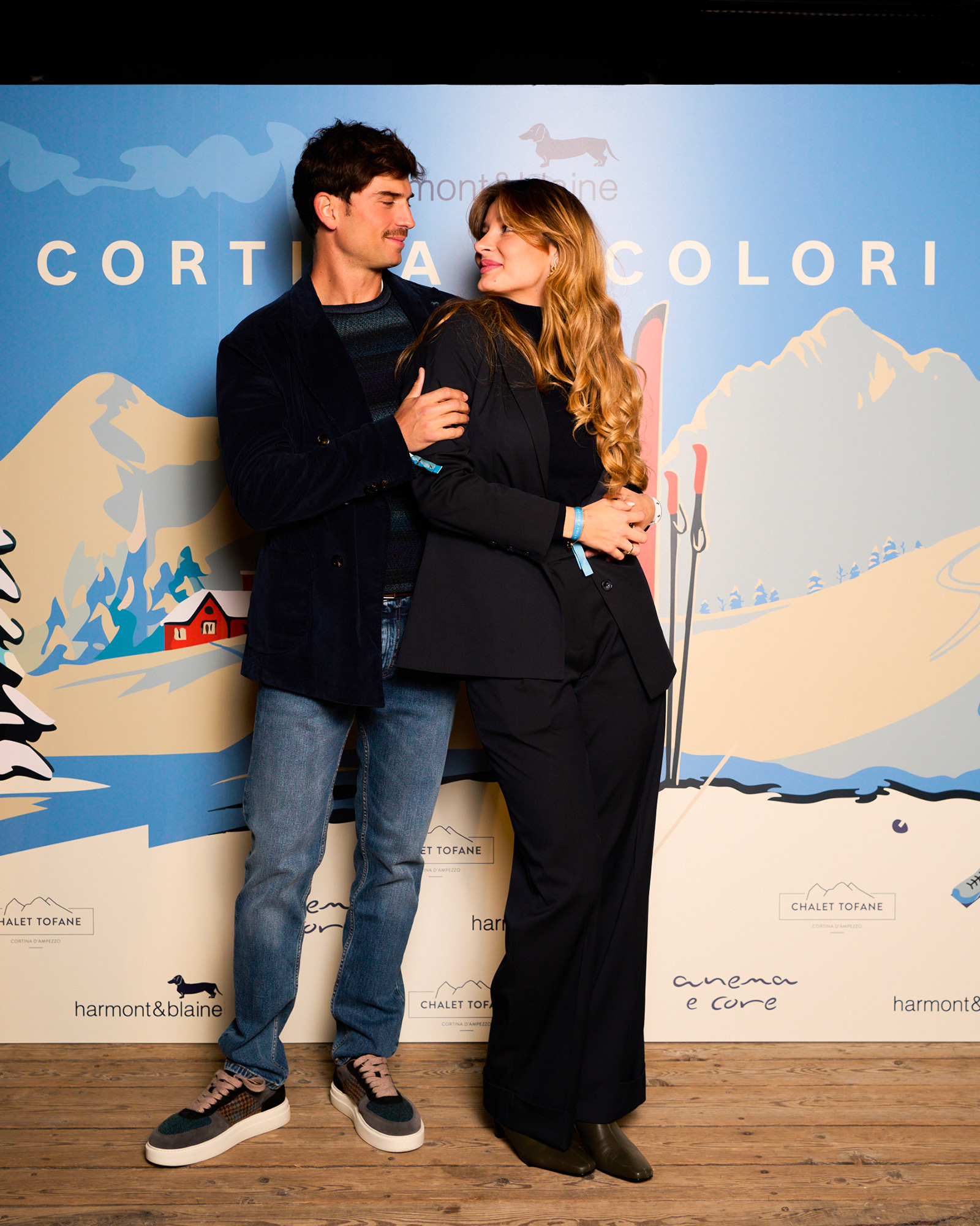 Couple in stylish outfits posing in front of the Cortina a Colori backdrop.