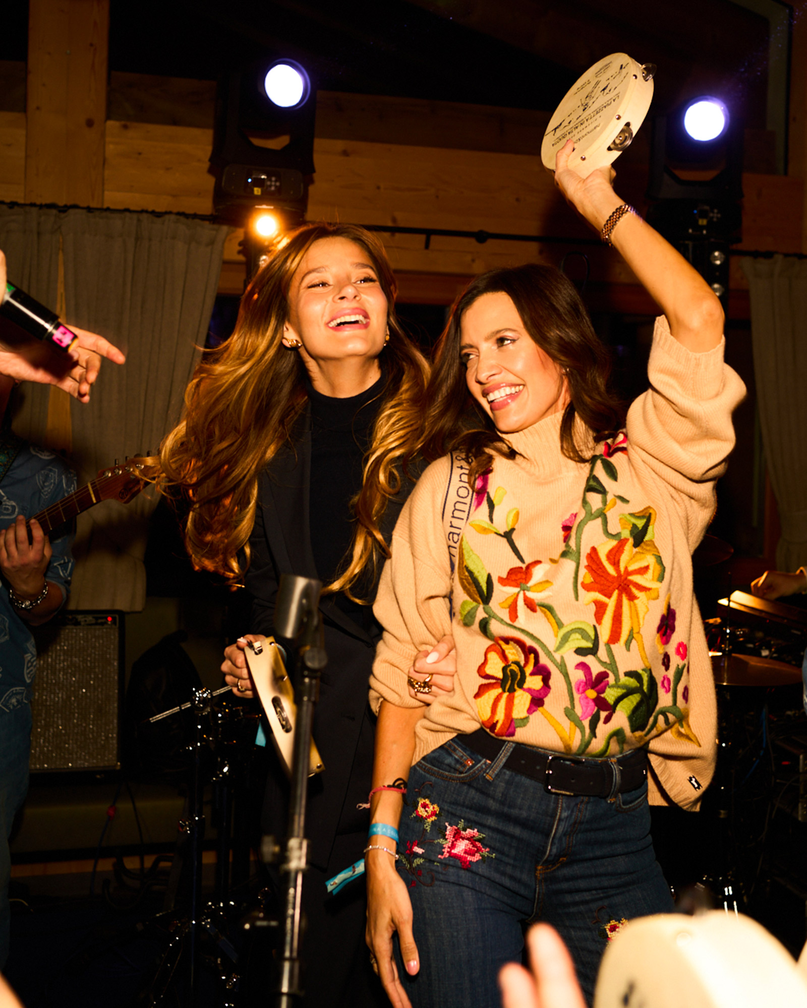 Two women dancing with tambourines, wearing a floral sweater and dark outfits.