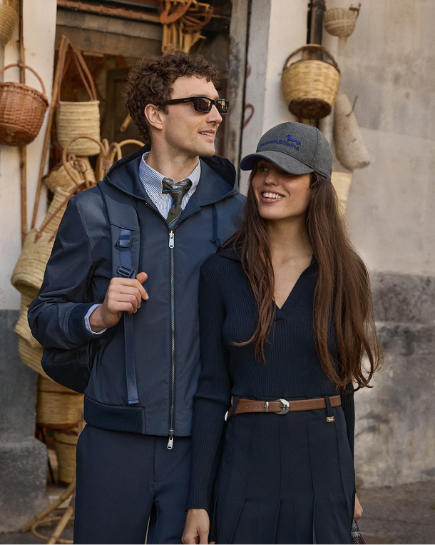 Immagine di campagna per la preview Autunno/Inverno 2026 di Harmont & Blaine, focus "Reinventing Sport". Una giovane coppia cammina sorridente accanto a una bottega di cestini in vimini: l'uomo indossa una giacca a vento blu scuro con cappuccio sopra camicia e cravatta, pantaloni scuri e occhiali da sole; la donna sfoggia un cappellino grigio con logo blu del brand, maglia e gonna a pieghe blu scuro con cintura in pelle marrone.