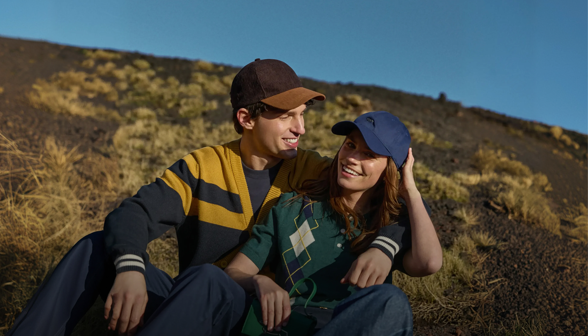 Questa &egrave; un'immagine di campagna per la preview autunno-inverno 2026 di Harmont & Blaine. L'immagine mostra una giovane coppia seduta vicina all'aperto su un pendio erboso e roccioso sotto un cielo blu terso. L'uomo &egrave; a sinistra e indossa un cardigan a coste a blocchi di colore senape e blu scuro con profili a contrasto sui polsini, un berretto bicolore marrone e marrone chiaro e pantaloni scuri. La donna &egrave; a destra, con un braccio intorno alle spalle dell'uomo, e indossa una polo a maniche corte verde con un motivo argyle geometrico color crema e blu scuro, e un cappellino da baseball blu scuro. Entrambi sorridono e guardano verso l'orizzonte, proiettando un'immagine di intimit&agrave; felice e rilassata.