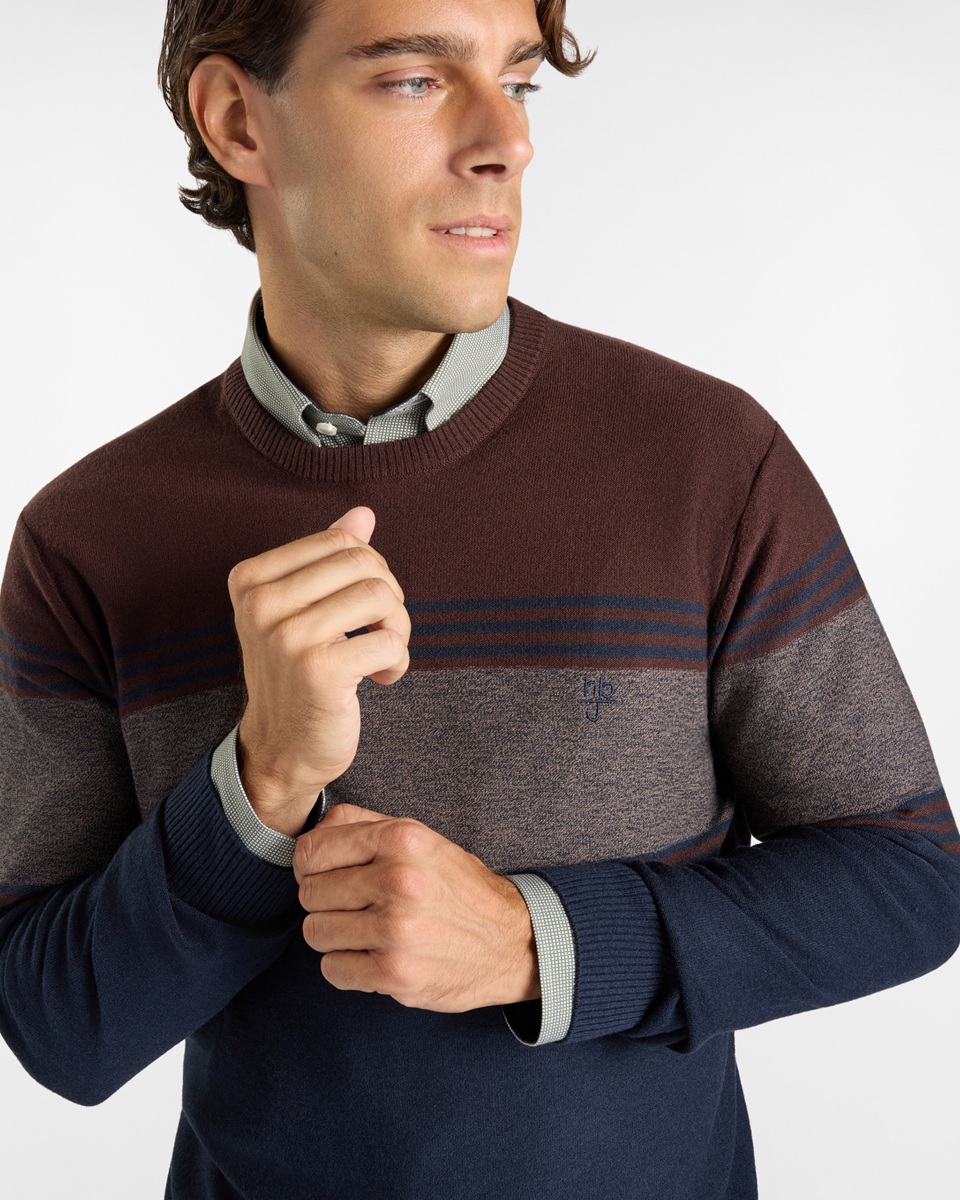 Uomo sorridente che indossa una giacca impermeabile blu scuro con cappuccio e un cappellino con visiera di colore bordeaux sotto il cappuccio.