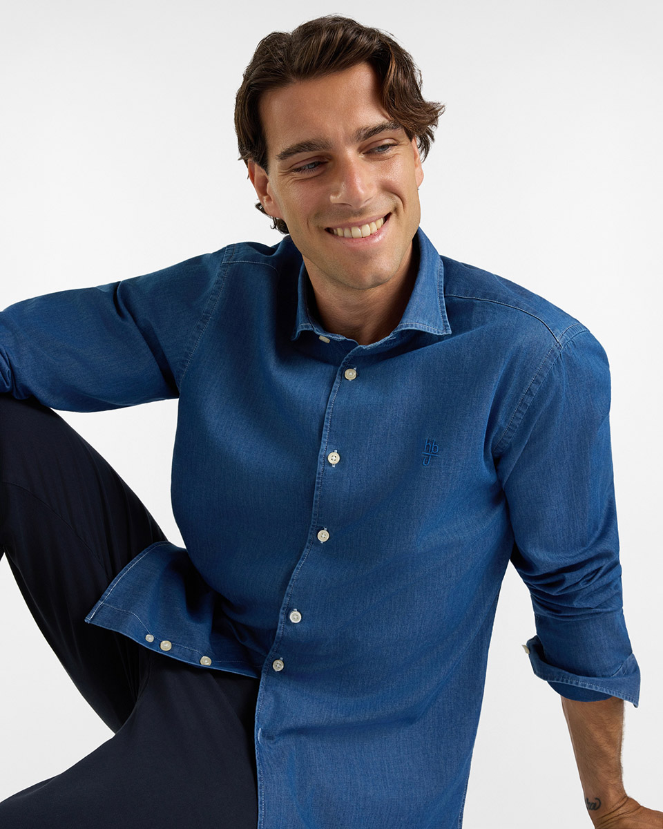 Per la categoria camicie HB Jeans, un modello sorridente indossa una camicia in denim blu medio a maniche lunghe risvoltate, con colletto francese, bottoni chiari a contrasto e logo ricamato tono su tono sul petto, abbinata a pantaloni scuri su sfondo bianco