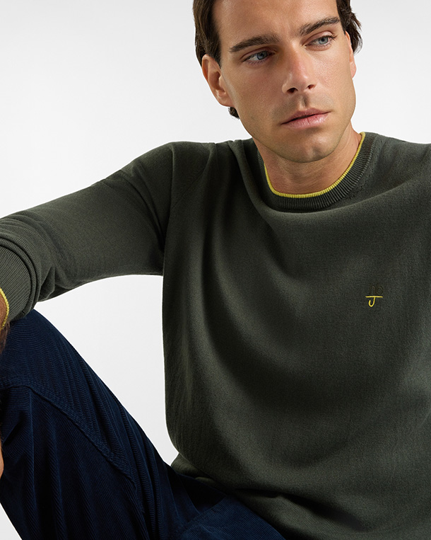 Per la categoria maglieria HB Jeans, un modello indossa un maglione girocollo verde militare a maniche lunghe, rifinito con un sottile profilo giallo a contrasto sul colletto e sui polsini, impreziosito da un piccolo logo ricamato in giallo sul petto e abbinato a pantaloni scuri su sfondo chiaro