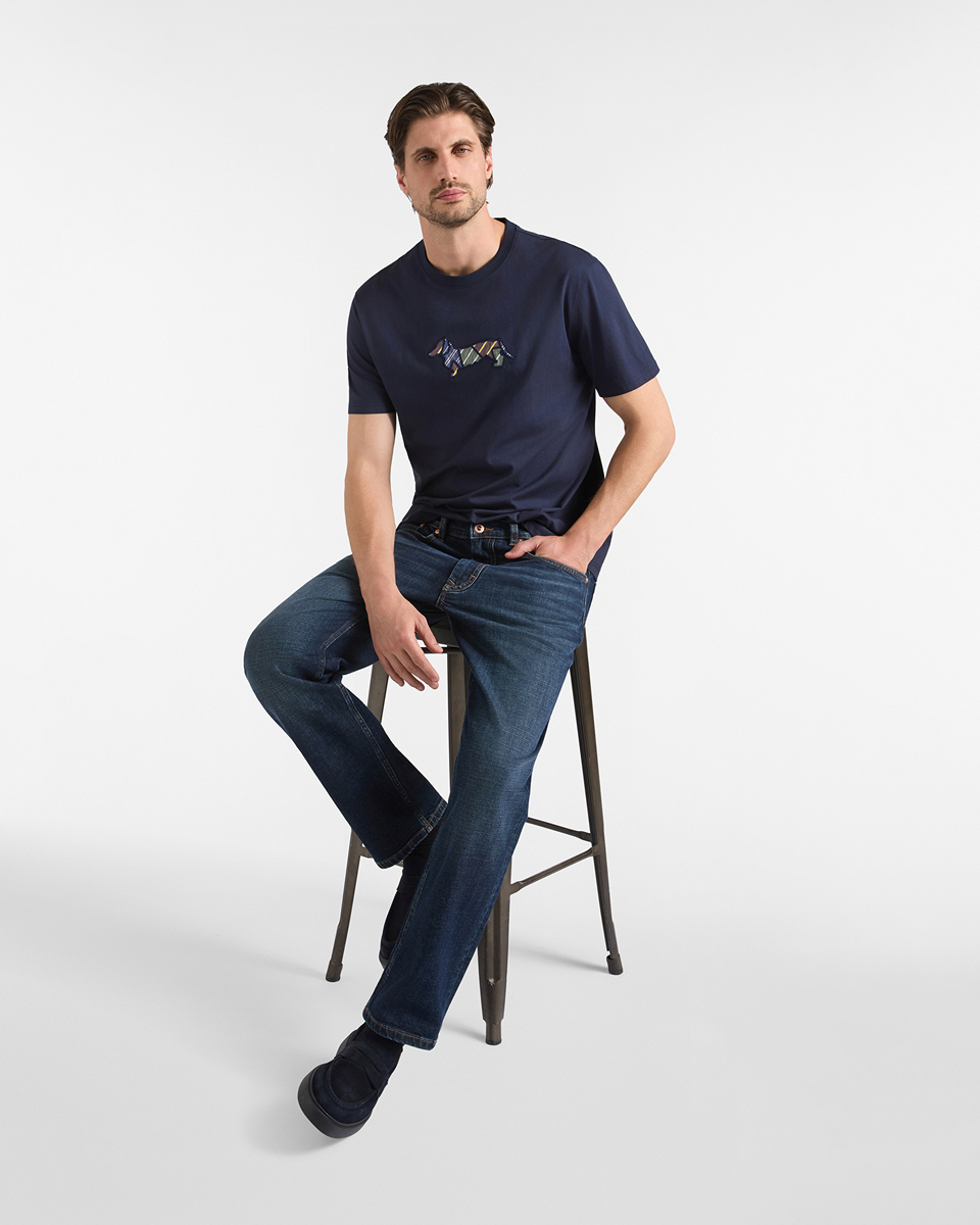 Un uomo castano seduto su uno sgabello alto in metallo, indossa una t-shirt blu scuro a maniche corte con logo ricamato sul petto, jeans blu scuro e mocassini in camoscio blu scuro.