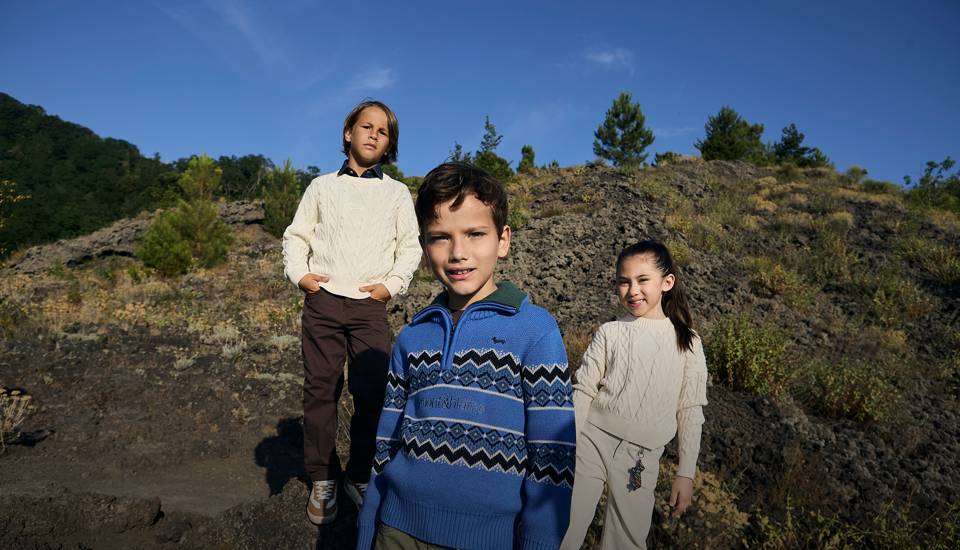 Three children are wearing Harmont&Blaine knitwear items&mdash;in the foreground, a girl in a light cable-knit sweater and a boy in a blue zip-up sweater with dark geometric patterns, while a third child in the background wears a light pullover&mdash;immersed in an outdoor natural setting under a clear blue sky.