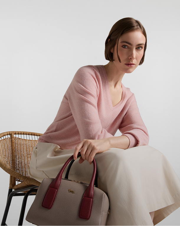 Donna seduta su una sedia in vimini che indossa un maglione rosa con scollo a V e gonna chiara, tenendo in mano una borsa bauletto beige con manici bordeaux e logo dorato.