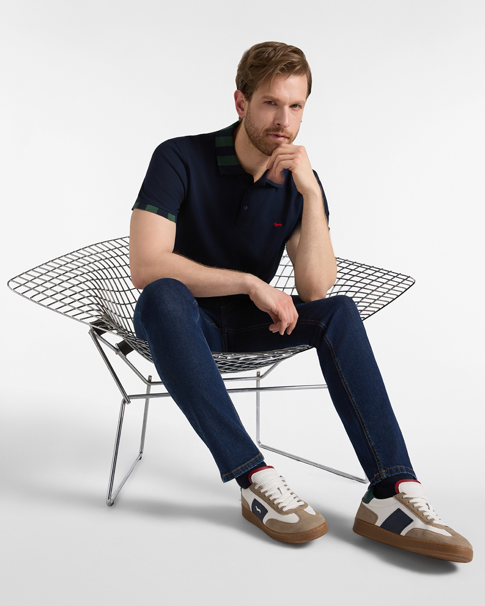 Un uomo biondo con la barba, seduto su una sedia in rete metallica cromata, indossa jeans scuri slim fit, polo blu con dettagli verdi e sneaker beige e blu.
