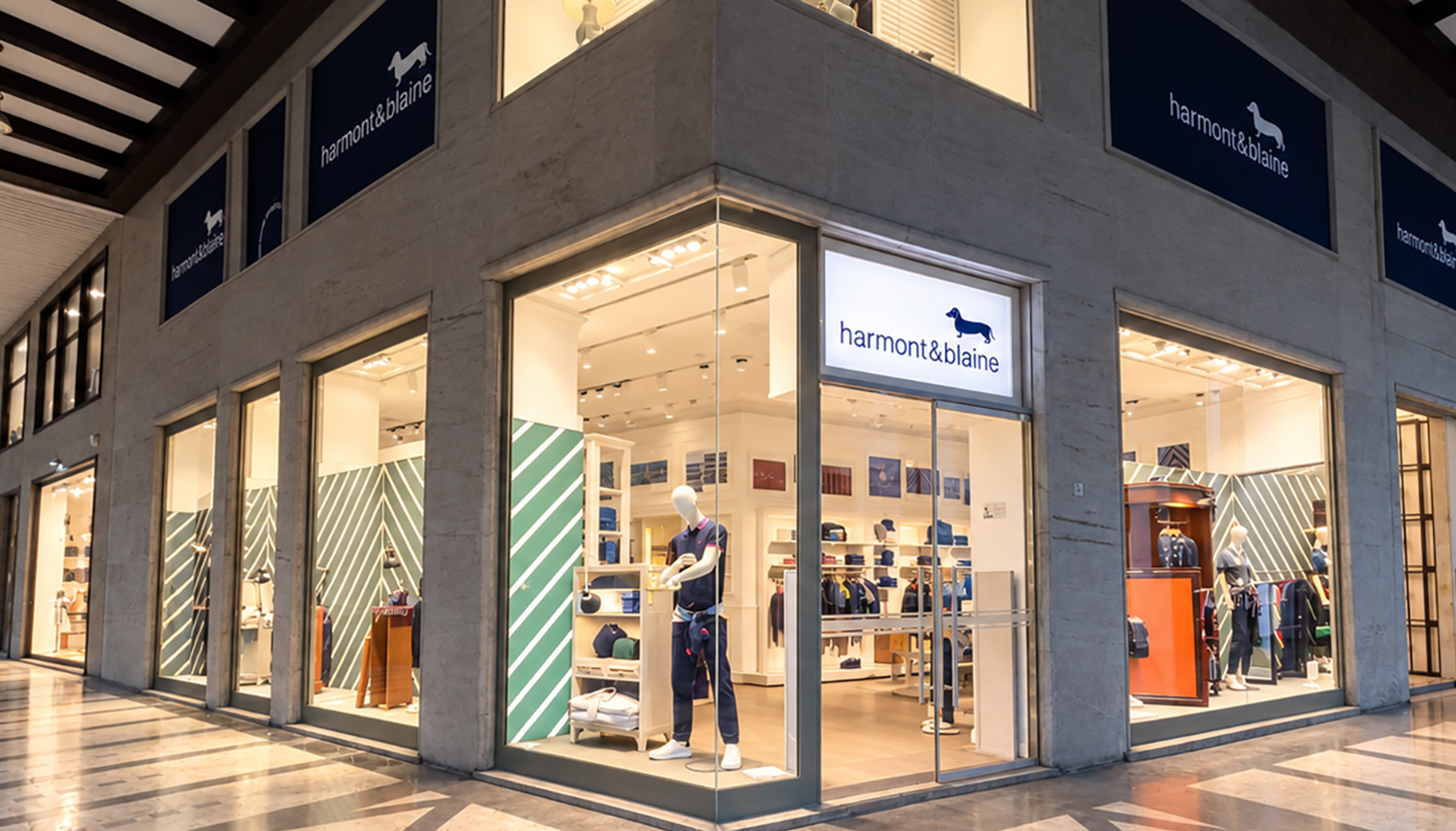 L'esterno di una boutique Harmont & Blaine situata in una galleria commerciale, caratterizzata da ampie vetrine ad angolo, un'illuminazione calda e arredi moderni con pareti a righe bianche e verdi e manichini in esposizione.
