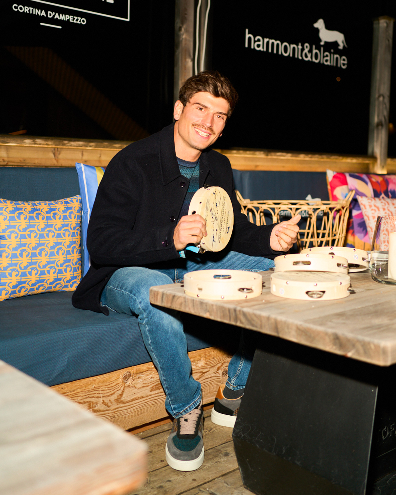 Man sitting with a tambourine, wearing a dark blue jacket, striped sweater, and jeans.