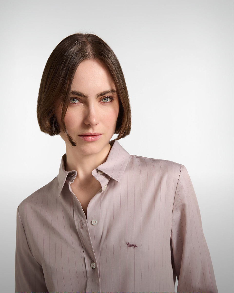 Donna con taglio di capelli bob che indossa una camicia gessata rosa antico con il logo del bassotto ricamato sul petto.