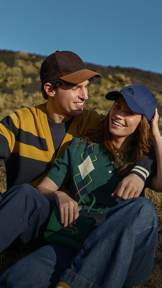 Questa &egrave; un'immagine di campagna per la preview autunno-inverno 2026 di Harmont & Blaine. L'immagine mostra una giovane coppia seduta vicina all'aperto su un pendio erboso e roccioso sotto un cielo blu terso. L'uomo &egrave; a sinistra e indossa un cardigan a coste a blocchi di colore senape e blu scuro con profili a contrasto sui polsini, un berretto bicolore marrone e marrone chiaro e pantaloni scuri. La donna &egrave; a destra, con un braccio intorno alle spalle dell'uomo, e indossa una polo a maniche corte verde con un motivo argyle geometrico color crema e blu scuro, e un cappellino da baseball blu scuro. Entrambi sorridono e guardano verso l'orizzonte, proiettando un'immagine di intimit&agrave; felice e rilassata.