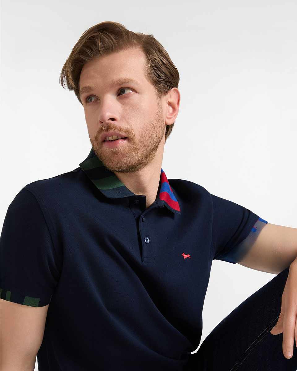 Immagine a mezzo busto, parte della campagna FW26 sul tema "Polo per Lui", ritrae un uomo con capelli biondo scuro e barba seduto e girato di tre quarti verso sinistra. Indossa una polo blu scuro con bottoni scuri e un colletto decorato a righe colorate in verde scuro, rosso scuro e viola, e il logo del marchio ricamato in rosso sul petto. Il fondo &egrave; bianco uniforme.